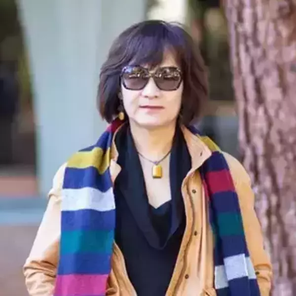 A woman with short dark hair, sunglasses, tan jacket, black top, and a colorful striped scarf with blue, yellow, white, red, green, pink bands. She has yellow earrings and an amber pendant.
