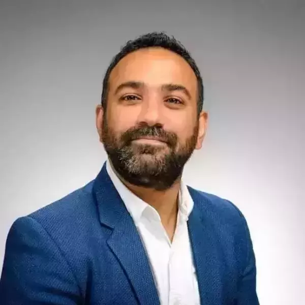 A man with dark hair and a beard smiles slightly, wearing a blue blazer over a white collared shirt, against a plain gray background.
